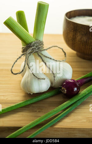 Three garlic bulbs tied up together with packthread on cutting board near onion and bowl full of salt. Unfocused background. Stock Photo