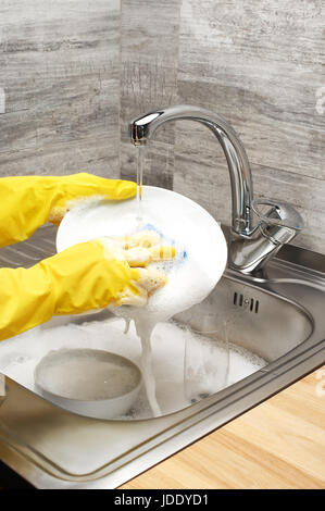 Close up of female hands in yellow protective rubber gloves washing white plate with blue rag against kitchen sink full of foam and tableware Stock Photo