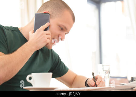 Handsome young man sitting at a table drinking coffee and reading the newspaper ads. Talking on a mobile phone and looking for a job for himself Stock Photo
