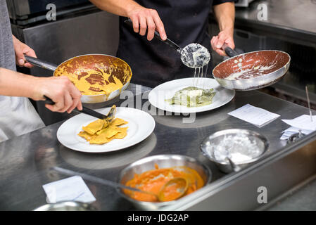 close-up of two unrecognizable chefs in aprons standing next to table in restaurant kitchen and serving ravioli pasta in plates from frying pans Stock Photo