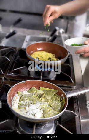 Close-up on ravioli in frying pan on gas-stove and incognito chef adding herbs to the second pasta dish on background Stock Photo