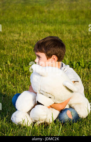 Lonely boy hugging teddy bear and sitting on the grass Stock Photo