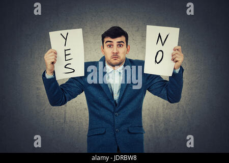 Confused young businessman holding two white papers in hands with yes and no text writed, isolated on grey wall background. Human emotion, face expres Stock Photo