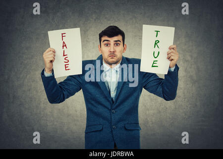 Confused young businessman holding two white papers try to choose between true or false, grey wall background. Human emotion, face expression, life pe Stock Photo