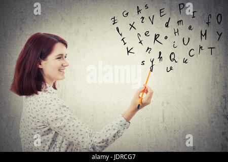 Young businesswoman, holding a pencil in her hand, drawing alphabet letters. Education and communication concept isolated on grey wall background. Stock Photo