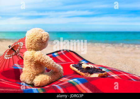 Teddy bear sitting at the beach with blanket, sunshade and sweet jam on bread roll to enjoy breakfast Stock Photo