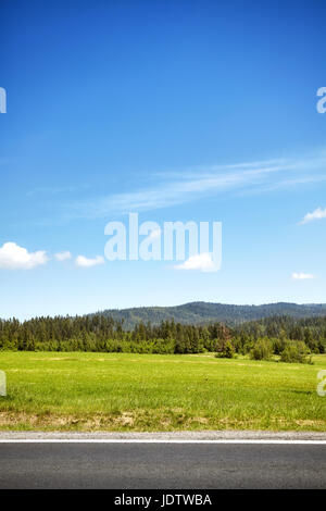 Green field by a road with blue sky and the mountain forest in the distance. Stock Photo