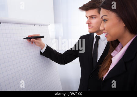 Businessman writing on flipchart with female colleague standing by him in office Stock Photo