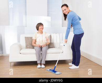 Portrait of caretaker cleaning floor with mop while senior woman sitting on sofa at nursing home Stock Photo