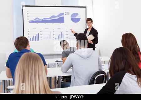 Young teacher teaching graphs drawn on whiteboard to college students in classroom Stock Photo