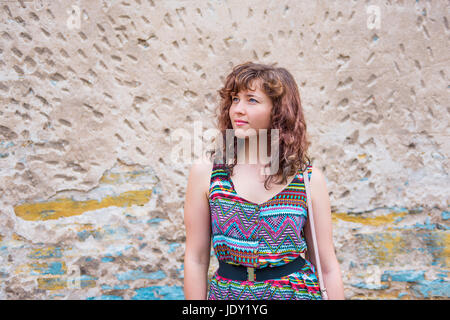 Young woman standing in front of aged stone wall background with rusty, peeling rocks and yellow and blue colors Stock Photo