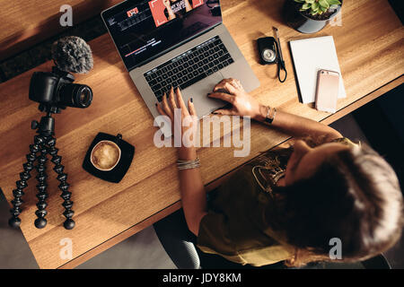 Top view of female vlogger editing video on laptop. Young woman working on computer with coffee and cameras on table.