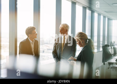 Three business colleagues standing behind the glass and discussing work. Corporate professional having a standing meeting in modern office. Stock Photo