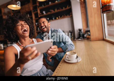 Happy man and woman having fun with phone while sitting at cafe. Couple enjoying at a coffee shop, sitting at table and laughing. Stock Photo