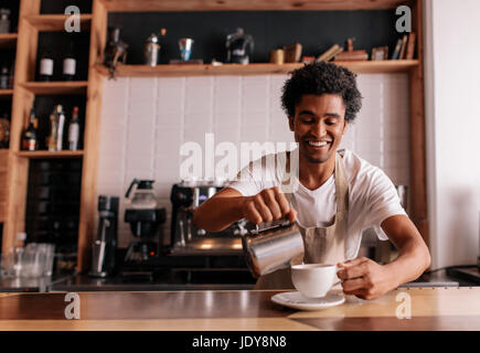 Professional barista making coffee on counter at cafe. Young african man pouring milk into cop and smiling. Stock Photo