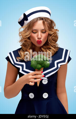 Creative retro photo of a fashionable pin-up sailor girl looking surprised with a broccoli ice cream in a waffle cone on blue background. Stock Photo