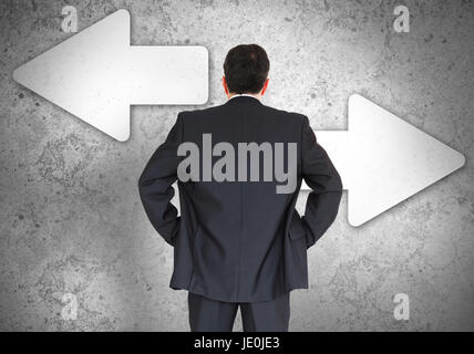 Businessman choosing which direction to go on grey wall background with white arrows Stock Photo