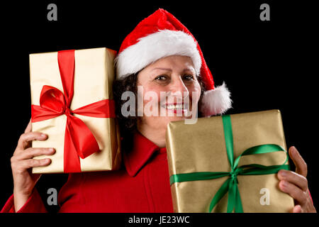 Perky senior woman is holding two gifts, one to her right cheek, the other in front of her chest. Both wrapped in plain golden paper with bows. Red Santa Claus cap. Isolated on black. December theme. Stock Photo