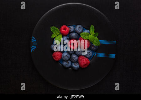 Fresh berries on black plate on black background, top view. Raspberries and blueberries with fresh mint leaf, minimal modern style. Stock Photo