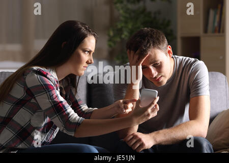 Girlfriend asking for explanation of her boyfriend sitting on a couch in the living room in a house interior with a dark background Stock Photo