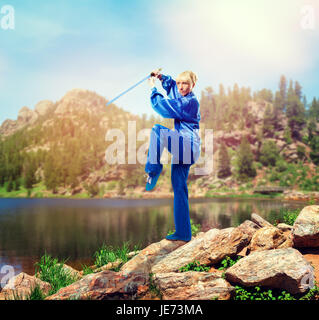 Female wushu master with sword against lake and mountains, martial arts. Woman in blue cloth on outdoor fight training Stock Photo