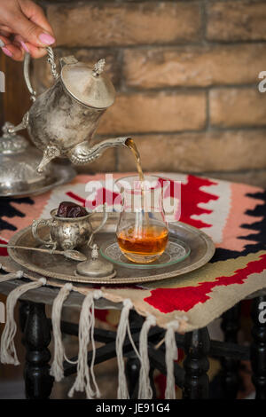 Tea set in oriental style in pear shaped glass with spoon and vintage kettle and dates fruit on silver tray on silver tray against brick wall Stock Photo