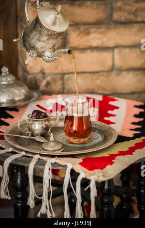 Tea set in oriental style in pear shaped glass with spoon and vintage kettle and dates fruit on silver tray on silver tray against brick wall Stock Photo