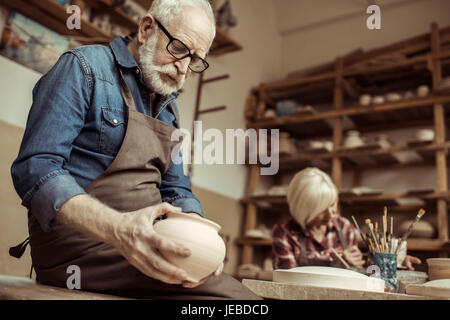 Senior potter in apron and eyeglasses examining ceramic bowl with woman working on background Stock Photo