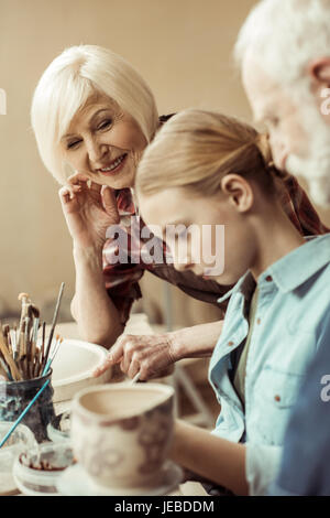 Side view of girl painting clay pot and grandparents helping at workshop Stock Photo