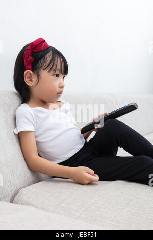 Asian Chinese little girl holding a TV remote control sitting on the sofa in the living room at home. Stock Photo