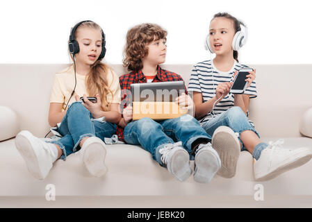 Cute kids using digital devices, listening music while sitting on couch Stock Photo