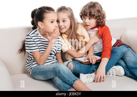 Two cute girls sharing secrets when sitting on couch Stock Photo