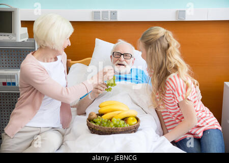 grandmother and granddaughter with fruits visiting patient in hospital. male patient in hospital bed concept Stock Photo