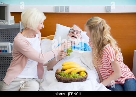 grandmother and granddaughter with fruits visiting patient in hospital. male patient in hospital bed concept Stock Photo