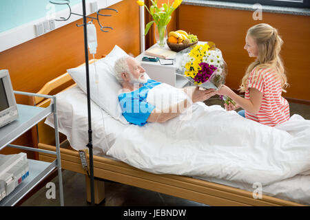 Smiling girl giving flowers to sick grandfather lying in hospital bed Stock Photo