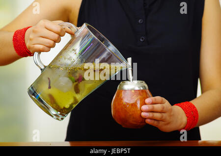 Woman holding trasnparent jar containing water and green herbs, pouring into traditional cup with typical metal straw sticking up, preparing popular d Stock Photo