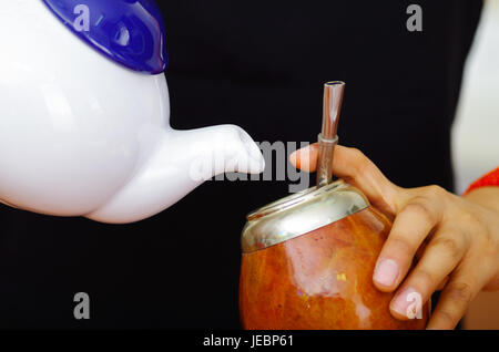 Woman holding white small jar pouring hot water into traditional cup with typical metal straw sticking up, preparing popular drink called mate. Stock Photo