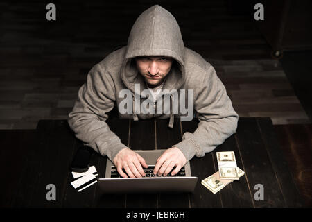 Young hacker in hoodie sitting at table and using laptop Stock Photo