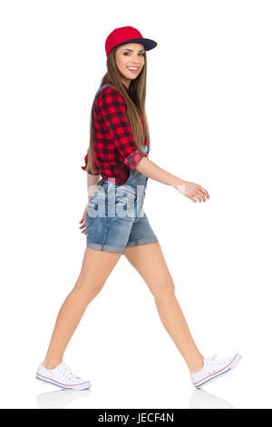 Woman in red lumberjack shirt, jeans shorts and white sneakers walking and looking at camera. Side view. Full length studio shot isolated on white. Stock Photo