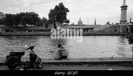 Romantic young couple embrace on the bank of the River Seine in Paris with their scooter nearby Stock Photo