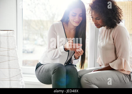 Two young attractive multiracial women friends checking a mobile phone for text messages as they sit close together on a high key windowsill with copy Stock Photo
