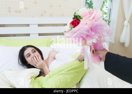 Picture showing Asian man surprise giving flowers and present to Asian young woman in bed at home. Surprise and anniversary concept Stock Photo