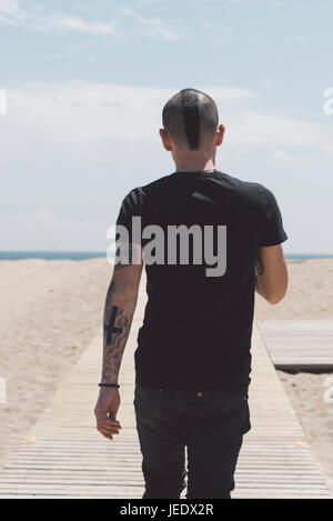 Back view of young man with mohawk haircut and tattoos walking on boardwalk to the beach Stock Photo