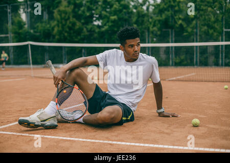 Exhausted male tennis player sitting on ground after tennis game Stock Photo