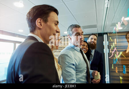 Multiracial business team developing a new project standing grouped around colorful sticky memos with ideas on a glass interface laughing and smiling Stock Photo