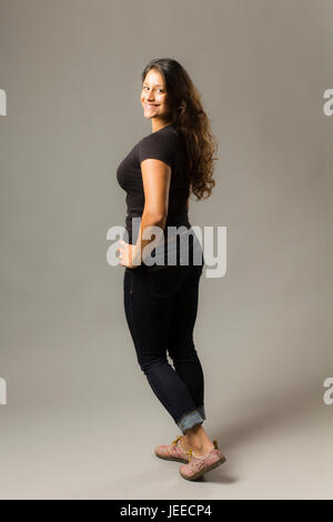 Young mixed race woman wearing a black t shirt and jeans stands looking over her shoulder while smiling at viewer Stock Photo