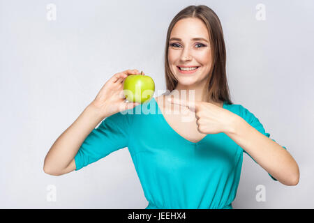 Young beautiful woman with freckles and green dress holding apple and pointing with finger. studio shot, isolated on light gray background. Stock Photo