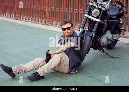 bearded redhead biker with beard in leather jacket sitting on floor near bike. Stock Photo