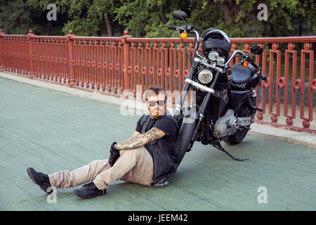 bearded redhead biker with beard in leather jacket sitting on floor near bike. Stock Photo