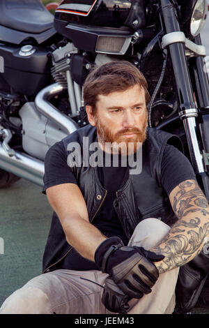 bearded redhead biker with beard in leather jacket sitting on floor near bike. Stock Photo